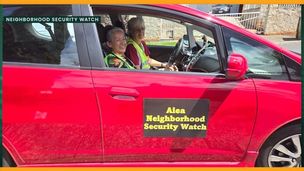 Two smiling volunteers in safety vests sit in a car marked Aiea Neighborhood Security Watch